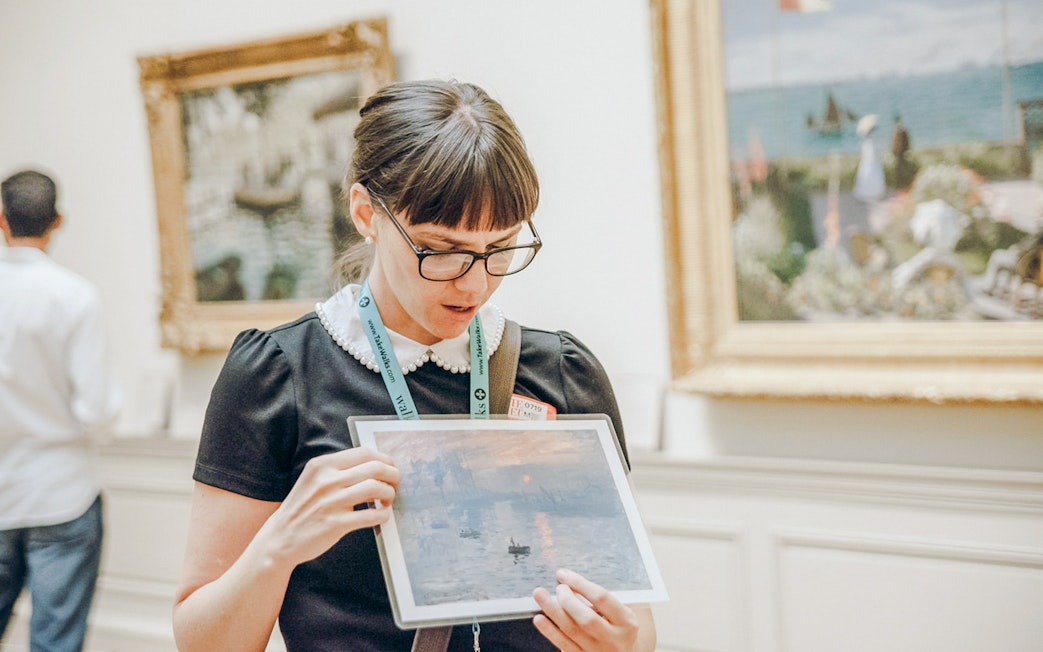 Tour guide at the Metropolitan Museum of Art holding a painting print in a gallery.