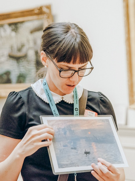 Tour guide at the Metropolitan Museum of Art holding a painting print in a gallery.