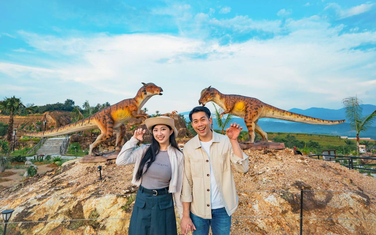 Couple posing with dinosaur sculptures in outdoor park setting.