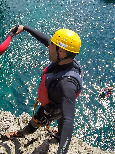 Coasteering participants in helmets and wetsuits preparing to jump from a rocky cliff into the sea.