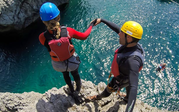 Coasteering participants in helmets and wetsuits preparing to jump from a rocky cliff into the sea.