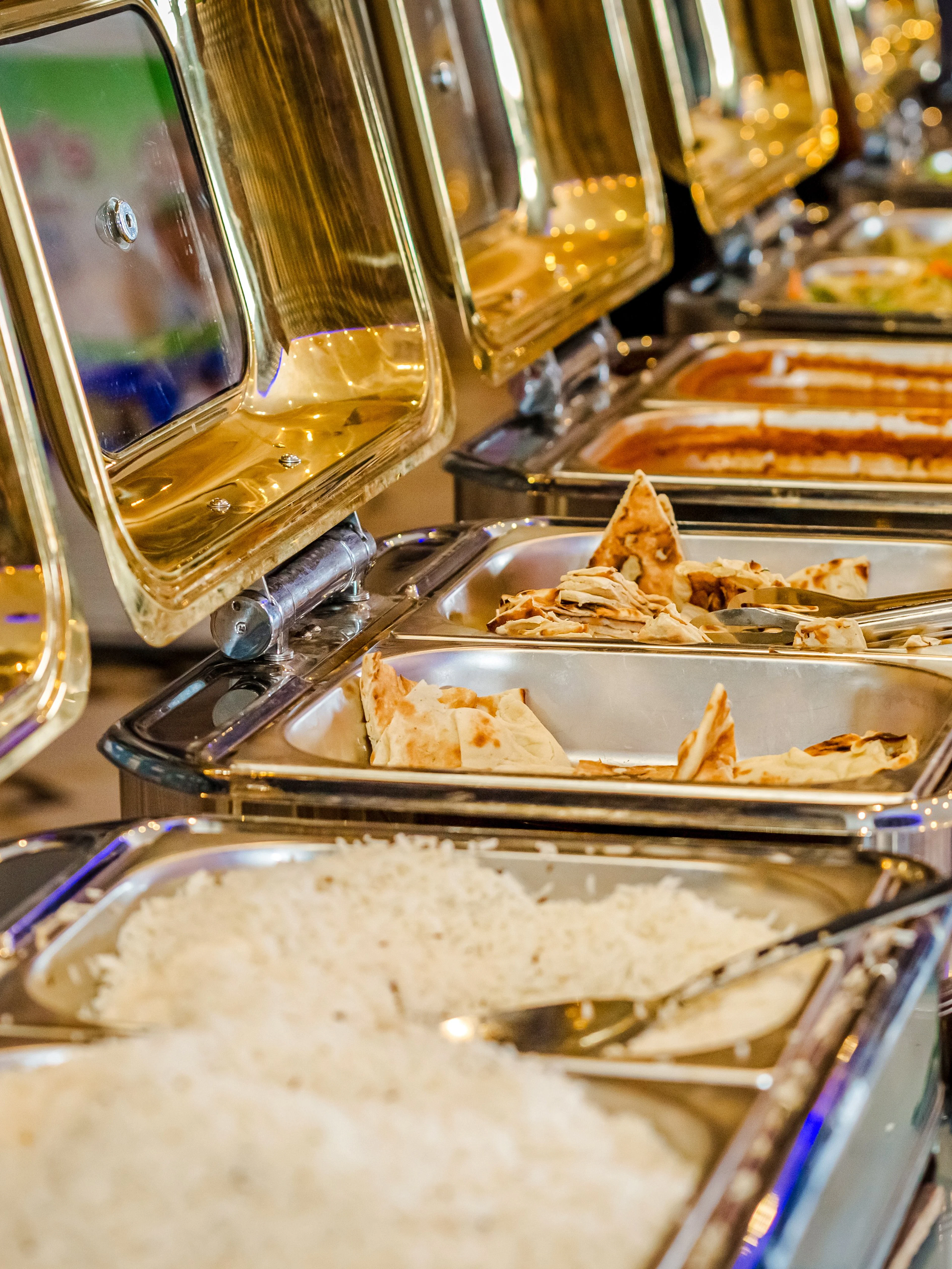 Indian buffet with rice, naan, and curry dishes in stainless steel trays.