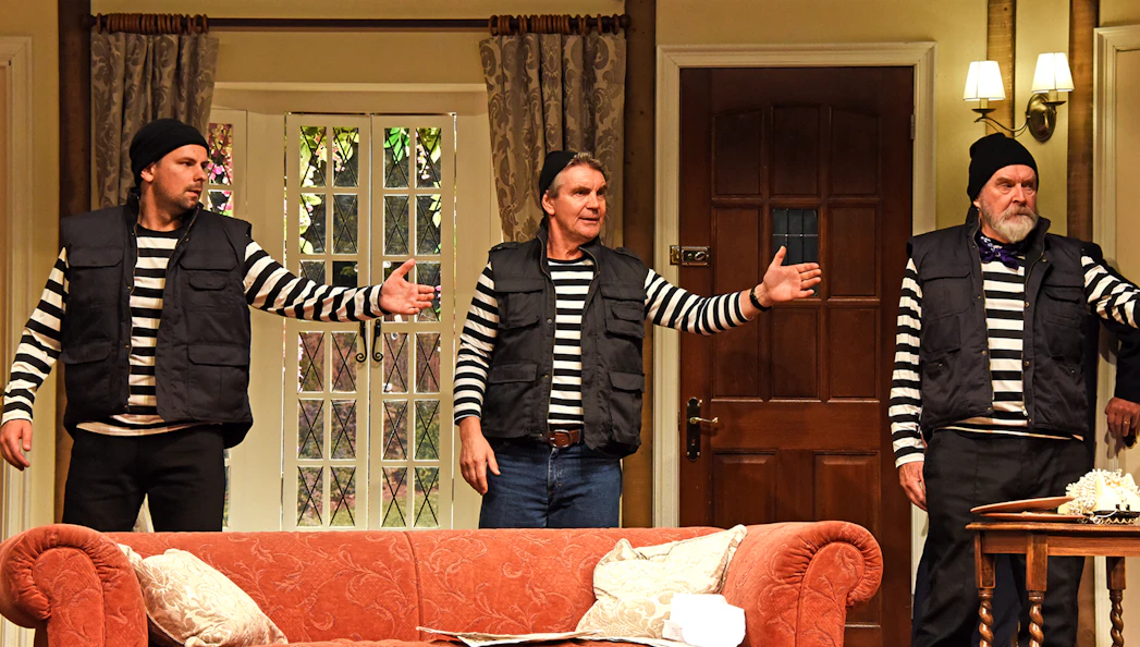 Three actors in striped shirts and vests perform a scene from "Noises Off" in a living room set.