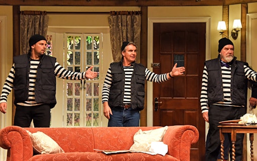 Three actors in striped shirts and vests perform a scene from "Noises Off" in a living room set.