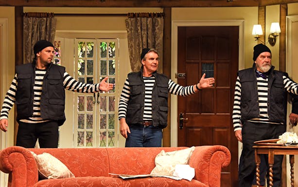 Three actors in striped shirts and vests perform a scene from "Noises Off" in a living room set.