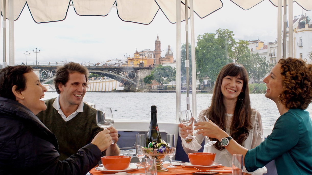 Tourists enjoying drinks and snacks on a Seville river cruise with Triana Bridge in the background.
