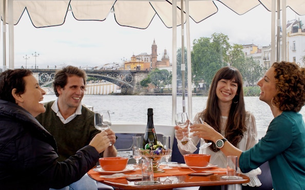 Tourists enjoying drinks and snacks on a Seville river cruise with Triana Bridge in the background.