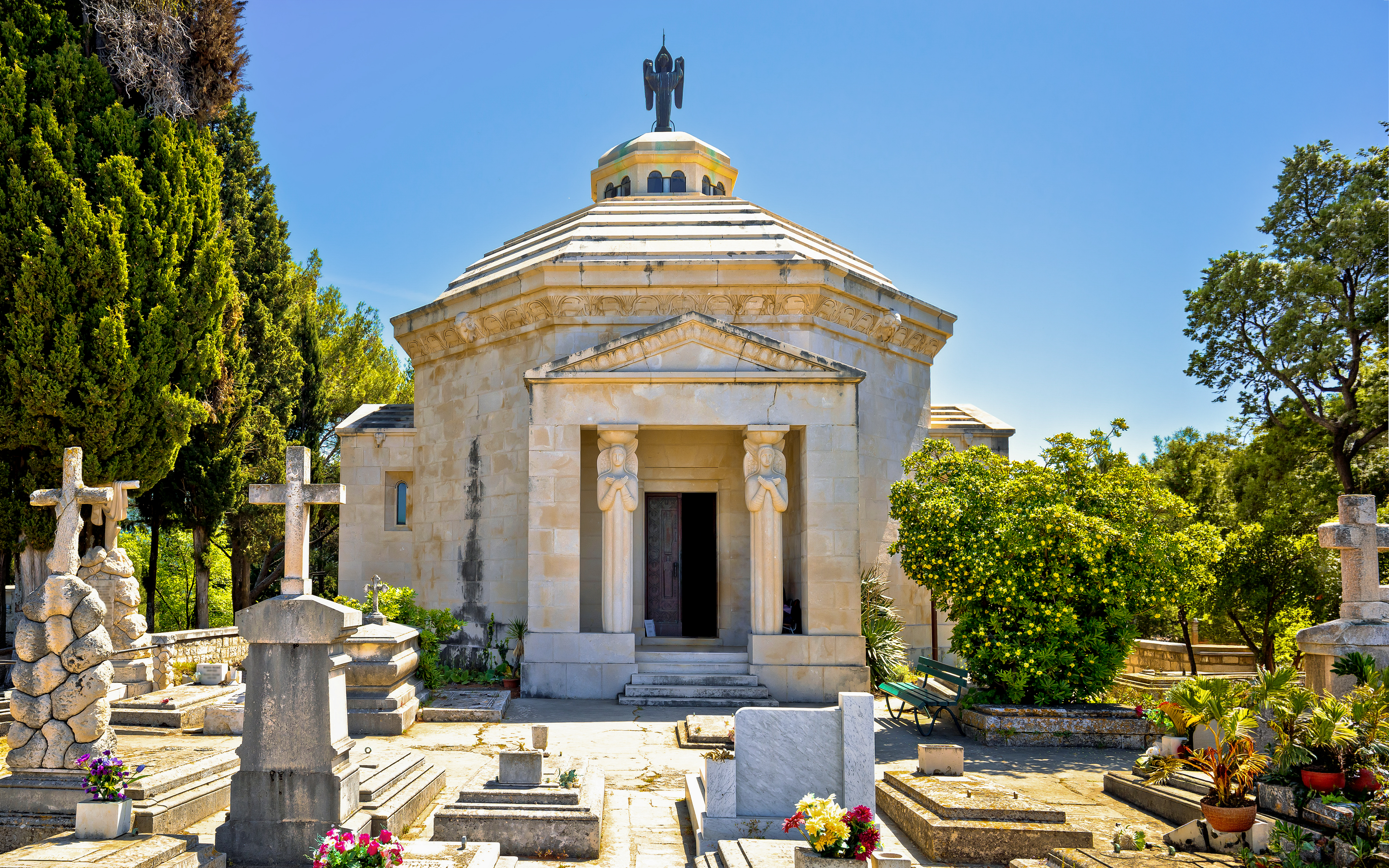 Racic Mausoleum in Cavtat graveyard surrounded by tombstones and greenery.