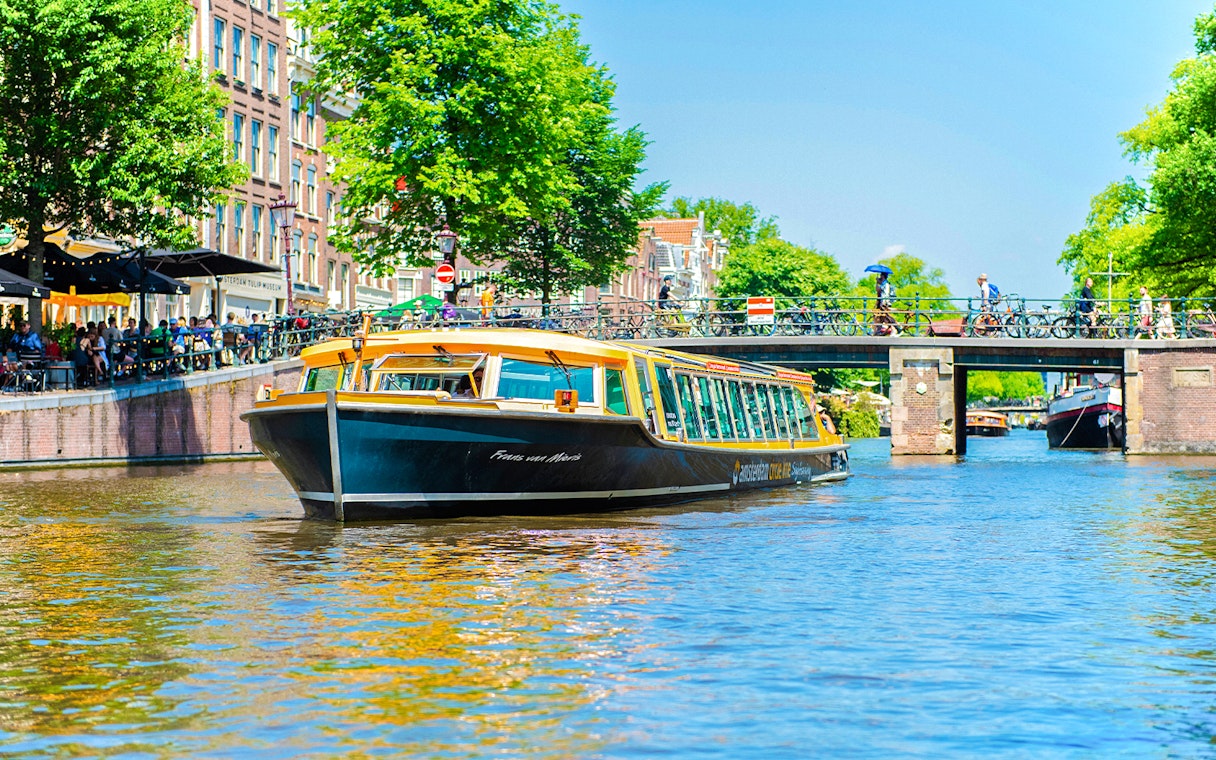 Canal boat cruising under a bridge in Amsterdam with people walking and cycling nearby.