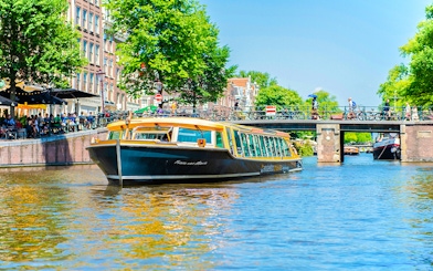 Canal boat cruising under a bridge in Amsterdam with people walking and cycling nearby.