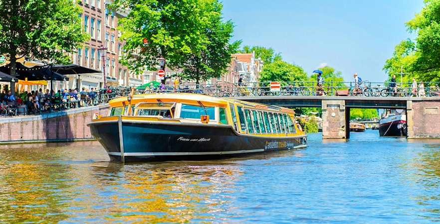 Canal boat cruising under a bridge in Amsterdam with people walking and cycling nearby.