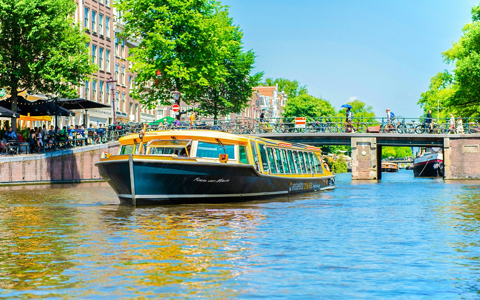Canal boat cruising under a bridge in Amsterdam with people walking and cycling nearby.