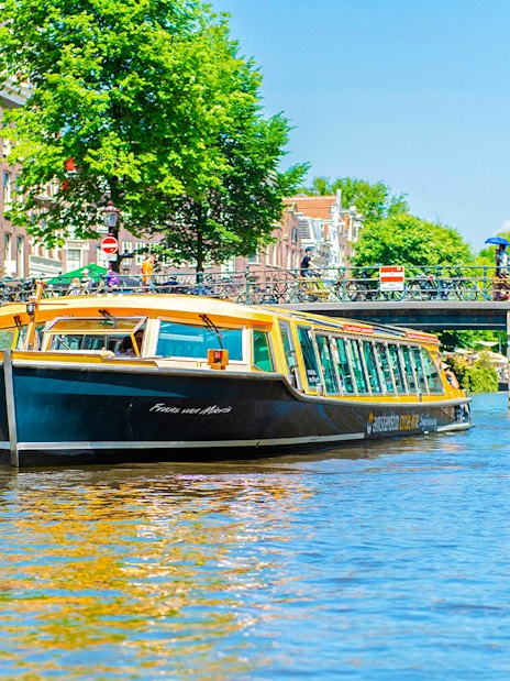 Canal boat cruising under a bridge in Amsterdam with people walking and cycling nearby.