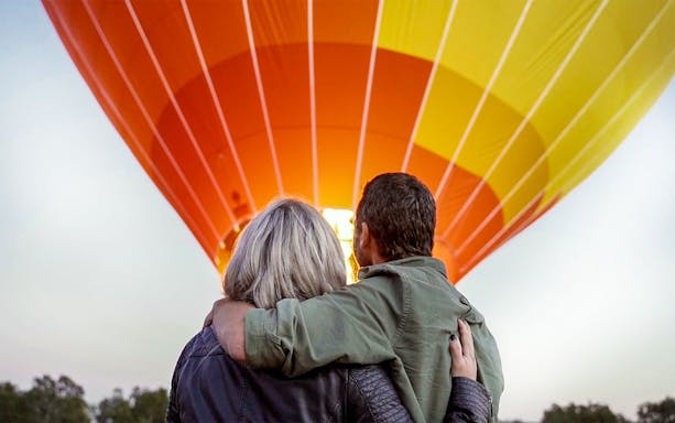 Couple embracing in front of a lit hot air balloon at sunrise.