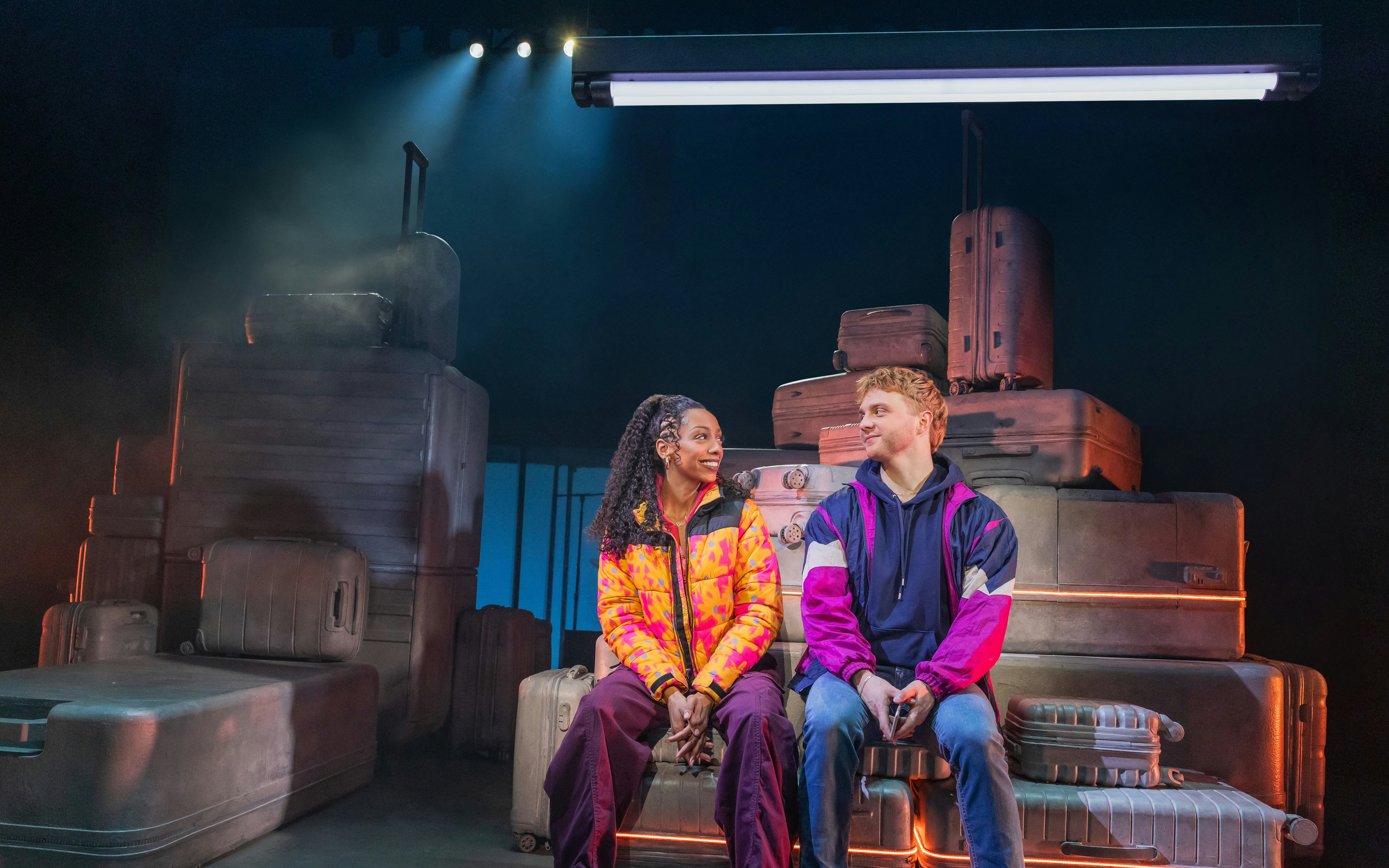 Two actors sitting on stage with stacked suitcases in "Two Strangers (Carry a Cake Across New York)" Broadway show.