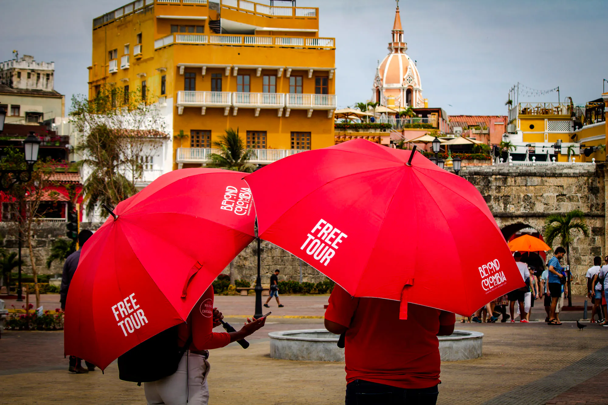 Tourists with red umbrellas in Cartagena's Walled Old City near historic buildings and dome.