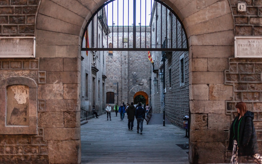 People walking through a historic stone archway in Barcelona's Raval district.