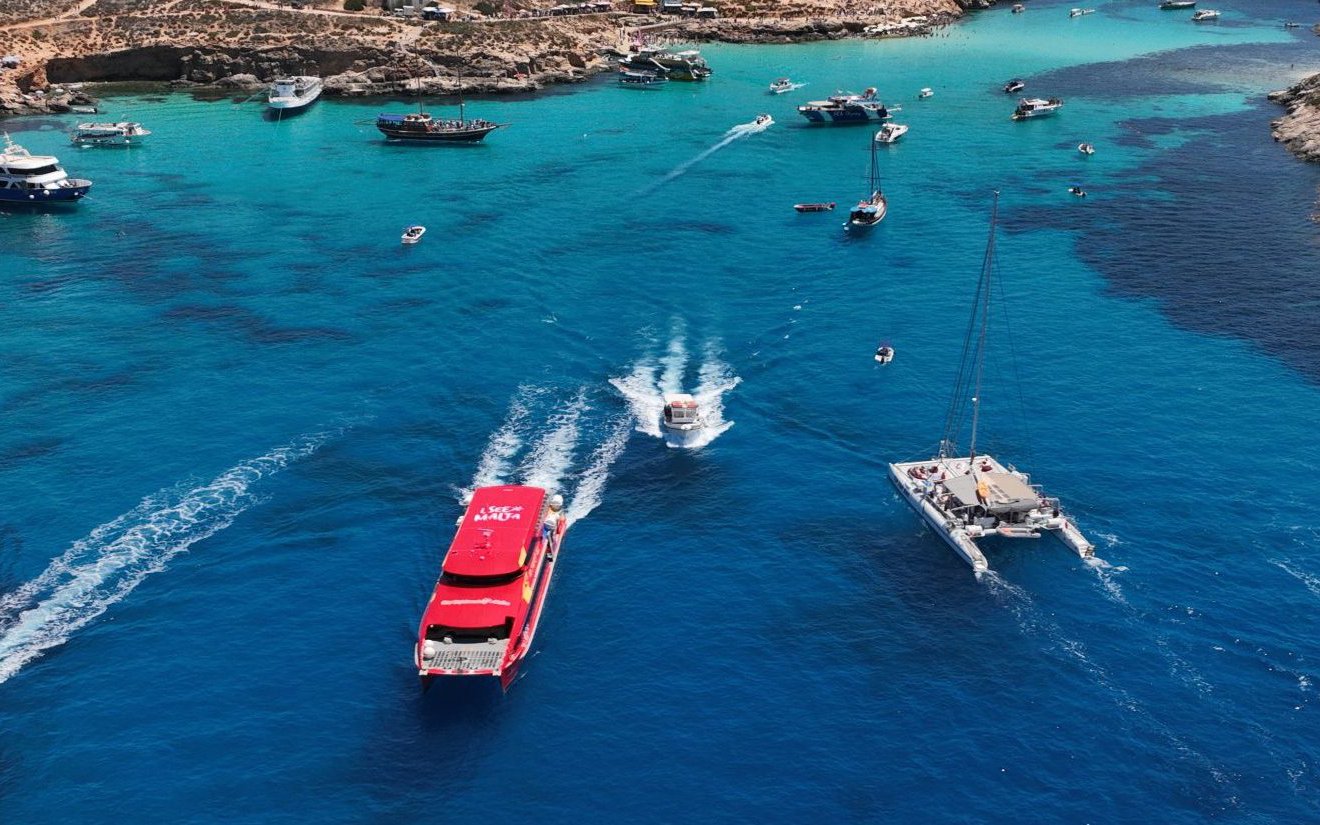 Boats cruising in a vibrant blue harbour with rocky coastline in the background.