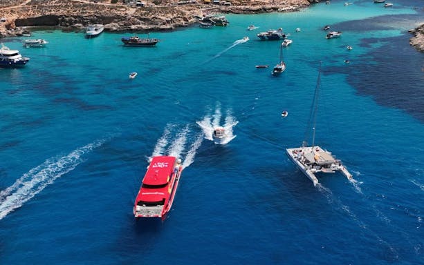 Boats cruising in a vibrant blue harbour with rocky coastline in the background.