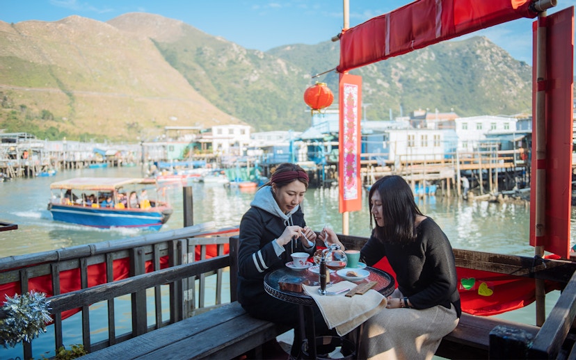 Two people dining at a waterfront café in Tai O, Hong Kong, with stilt houses and boats in the background.