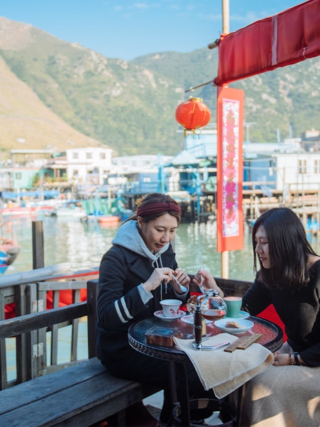Two people dining at a waterfront café in Tai O, Hong Kong, with stilt houses and boats in the background.