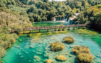 Visitors on a bridge over turquoise water at Krka National Park, Croatia, with waterfalls in the background.