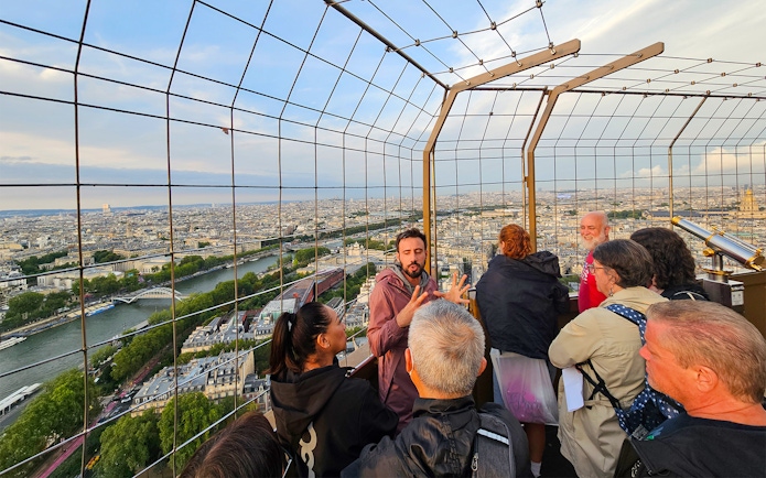 Tourists listening to guide on second floor of Eiffel Tower with Paris skyline.