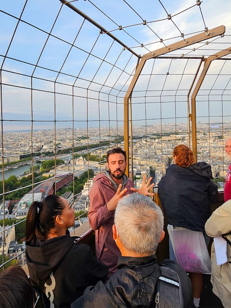 Tourists listening to guide on second floor of Eiffel Tower with Paris skyline.
