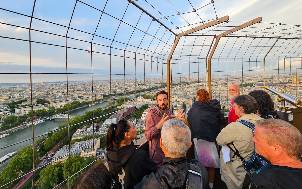 Tourists listening to guide on second floor of Eiffel Tower with Paris skyline.