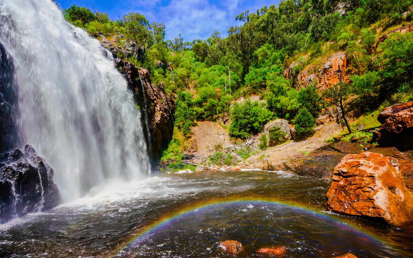 Waterfall cascading over rocks with a rainbow at MacKenzie Falls, surrounded by lush greenery.