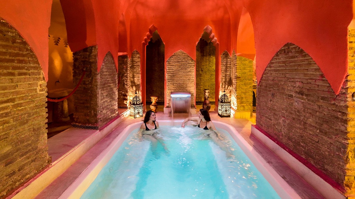 Two women relaxing in a Hammam pool in Granada, surrounded by traditional Arabic architecture.