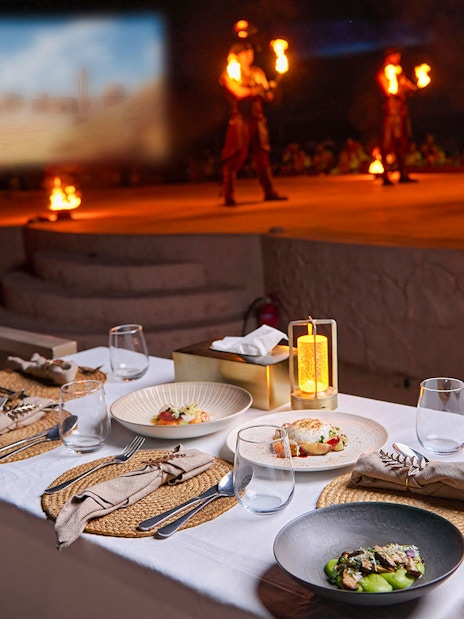 Dining setup with gourmet dishes at Noble Camp Desert Safari, fire performers in the background.