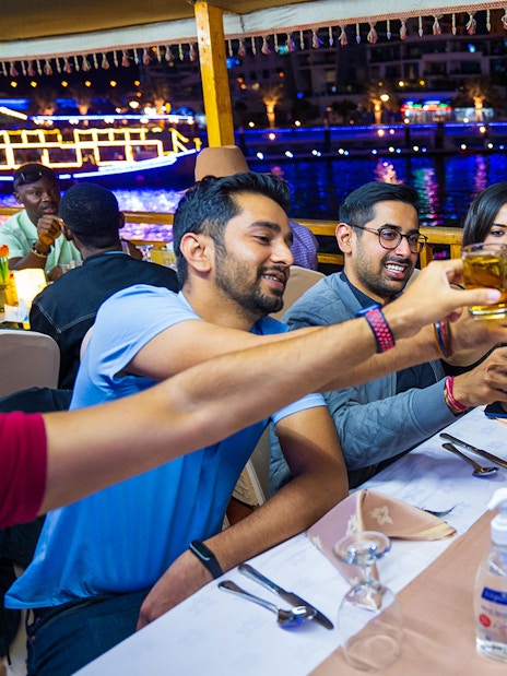 Guests toasting drinks on a Dhow Dinner Cruise in Dubai Creek at night.