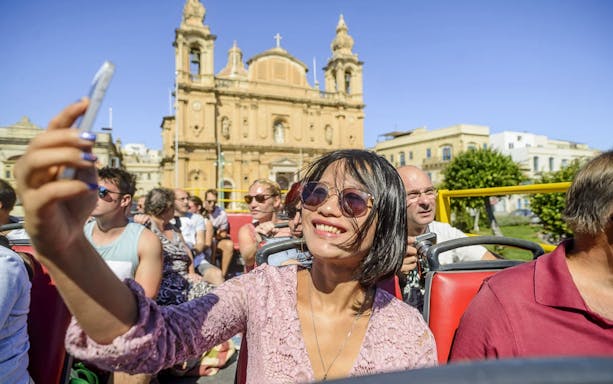 Tourist taking photos on Malta Hop-On Hop-Off Bus Tour with historic church in background.