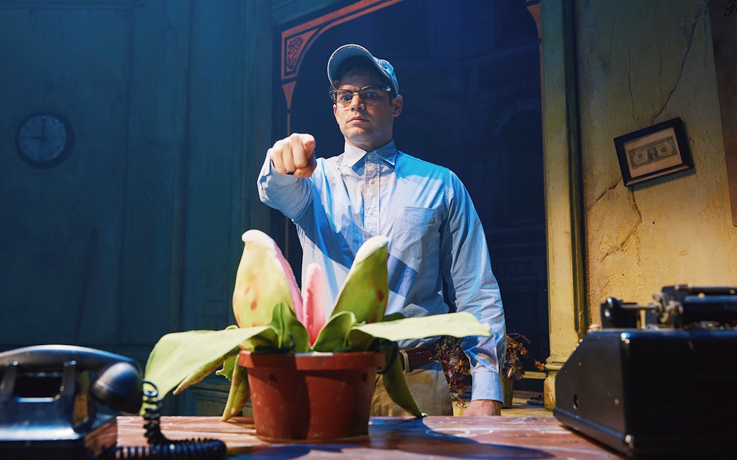 Man in blue shirt pointing at a plant in a scene from Little Shop of Horrors.