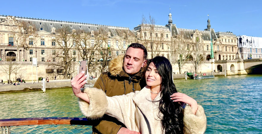 Couple taking a selfie on a Seine River cruise with Parisian architecture in the background.