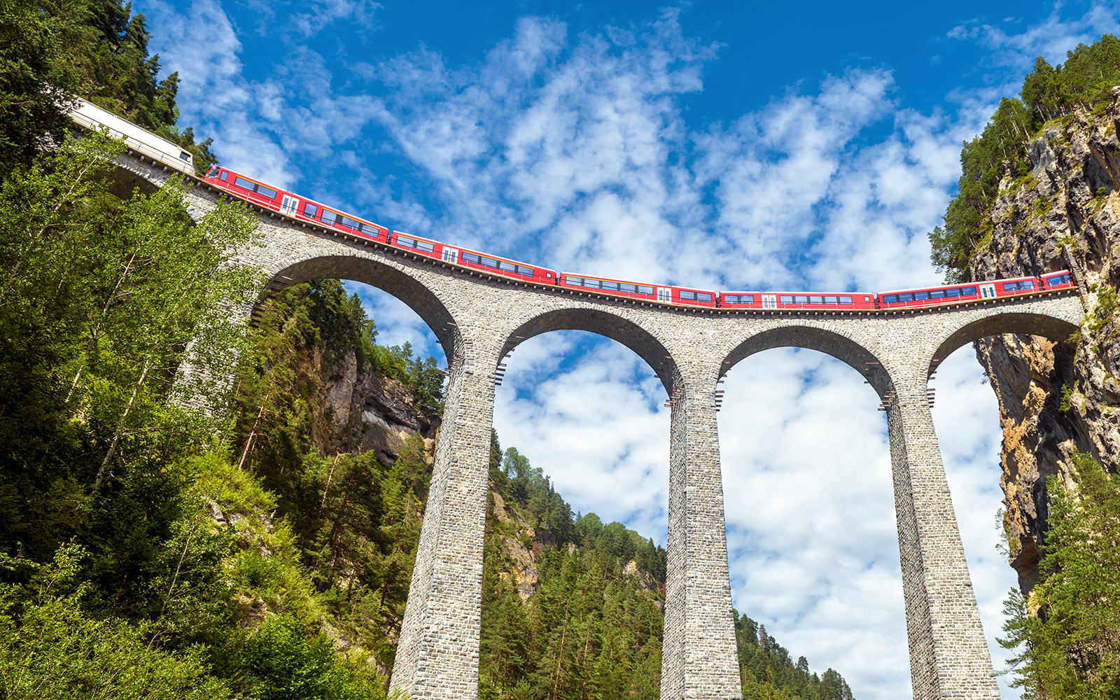 Glacier Express train runs on railroad bridge in Alpine mountains with scenic view of famous Swiss railway in summer.