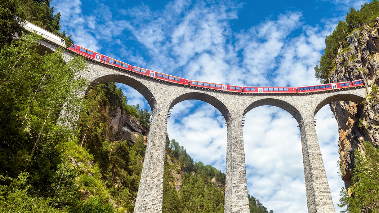 Glacier Express train runs on railroad bridge in Alpine mountains with scenic view of famous Swiss railway in summer.
