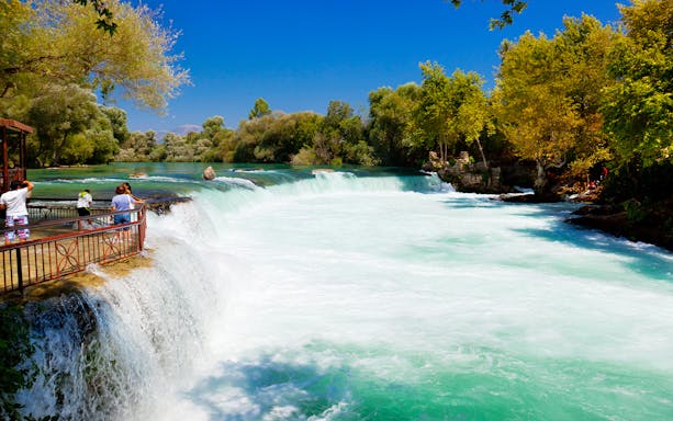 Manavgat River waterfall with tourists viewing from a platform, surrounded by lush greenery.