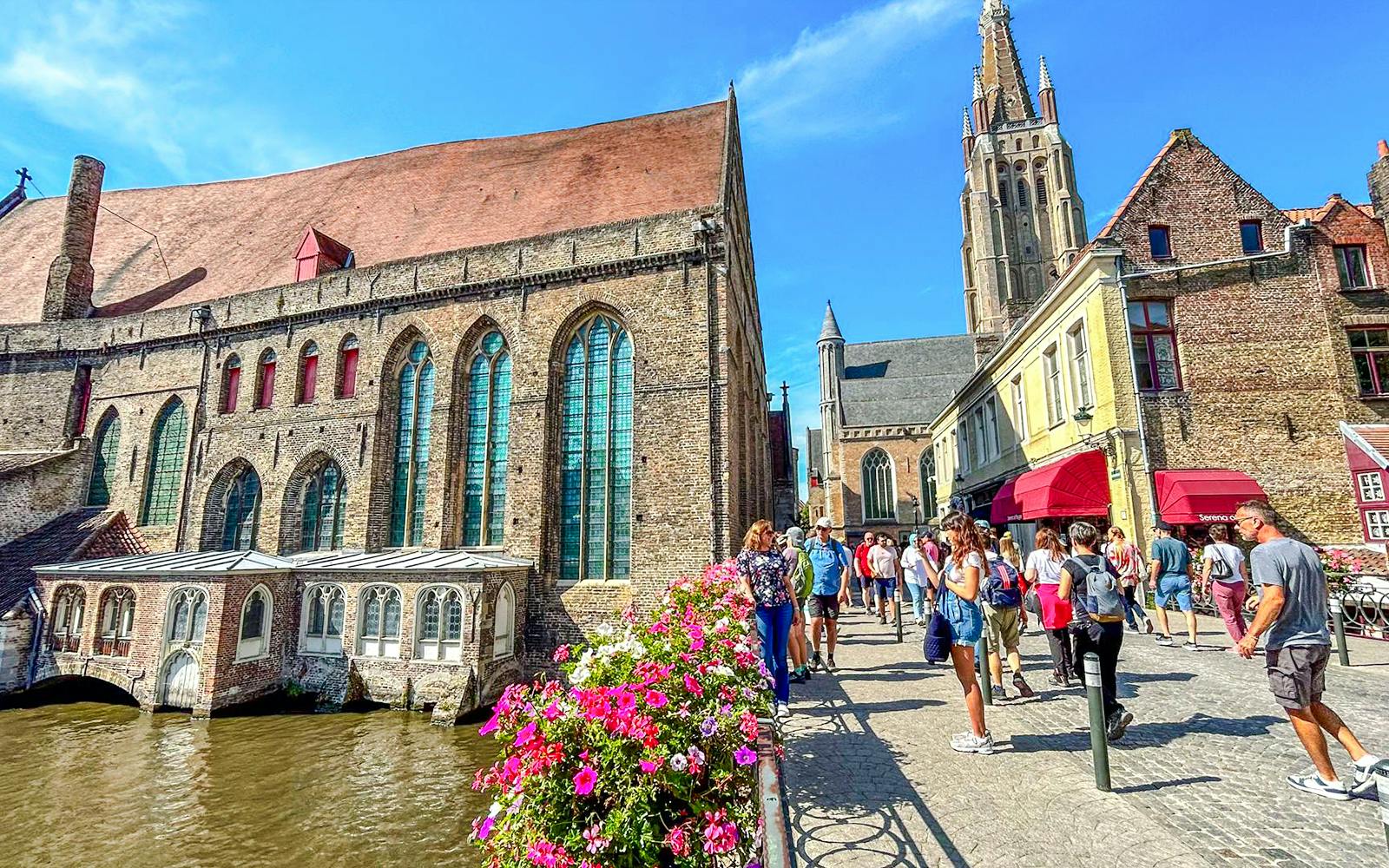 Old St. John's Hospital by a canal in Bruges, Belgium, with tourists walking nearby.