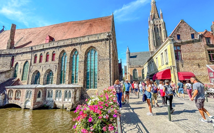 Old St. John's Hospital by a canal in Bruges, Belgium, with tourists walking nearby.