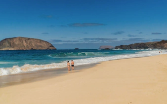 Couple walking on Las Conchas beach with ocean waves and rocky islands in the background.