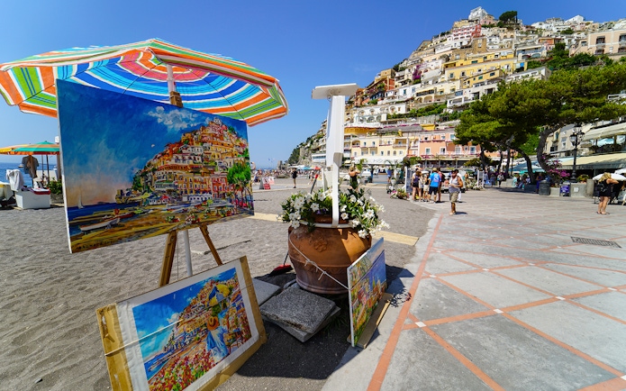 Colorful paintings of Positano displayed on a beach promenade, with hillside houses in the background.