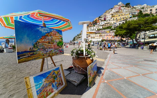 Colorful paintings of Positano displayed on a beach promenade, with hillside houses in the background.