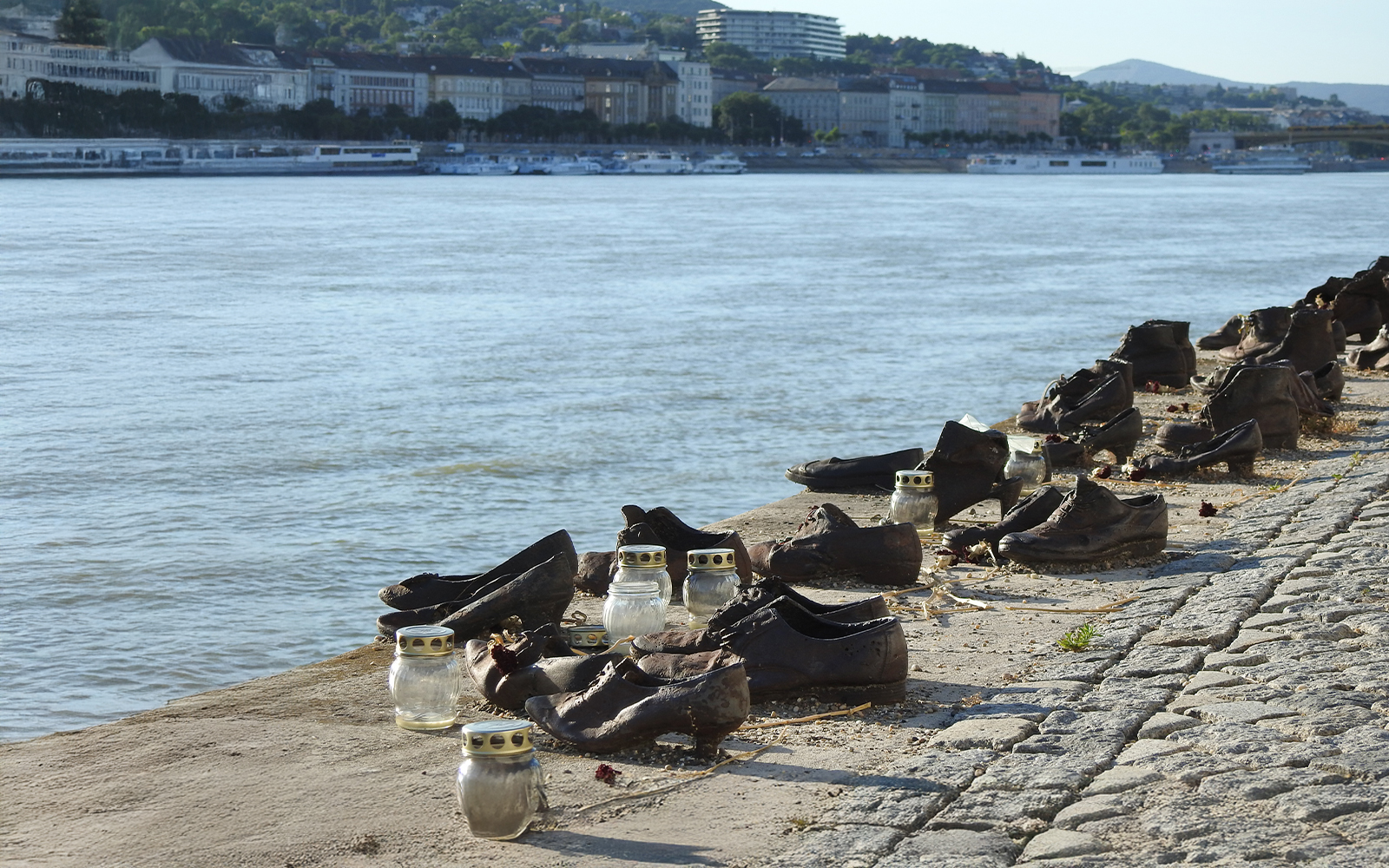 Shoes Memorial on the Danube Riverbank in Budapest, Hungary, with candles and cityscape in background.