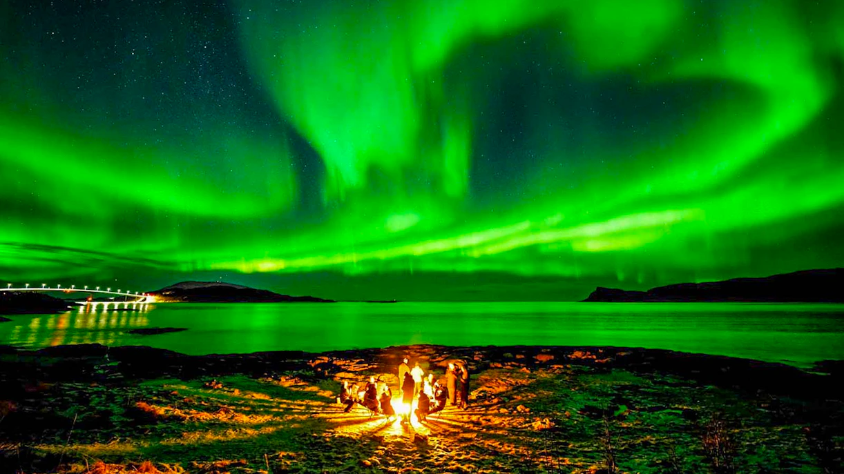 Guests gathered around a campfire under Northern Lights near Tromsø, Norway.