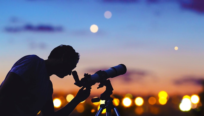 Astronomer using telescope to observe stars and Moon in a clear night sky.