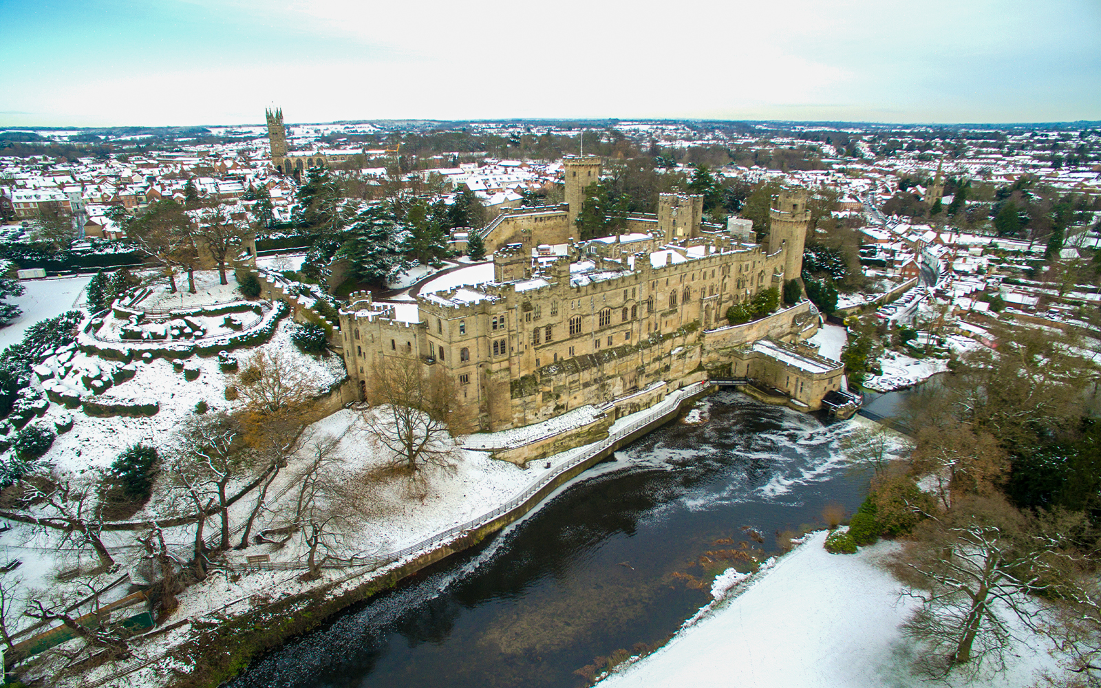 Warwick Castle covered in snow during Christmas, with surrounding snowy landscape and river.