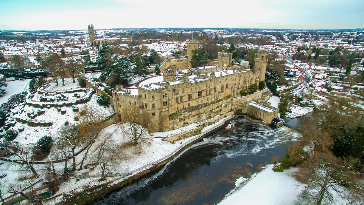 Warwick Castle covered in snow during Christmas, with surrounding snowy landscape and river.