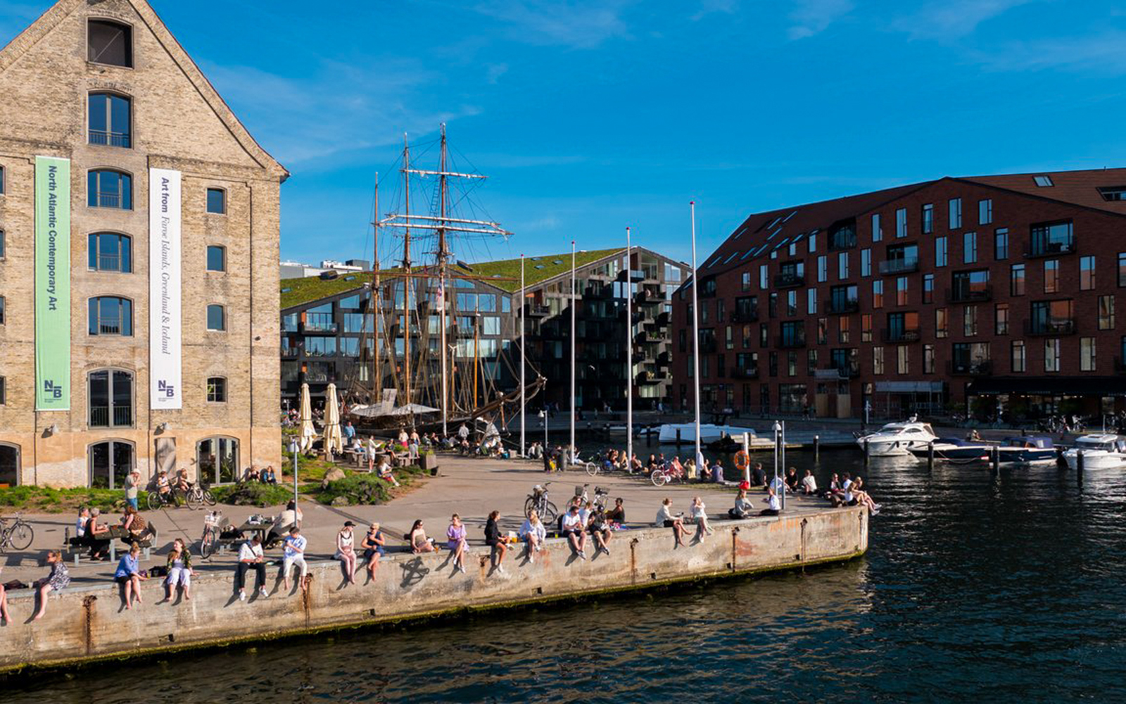 People relaxing by the waterfront in Copenhagen with historic buildings and boats in view.